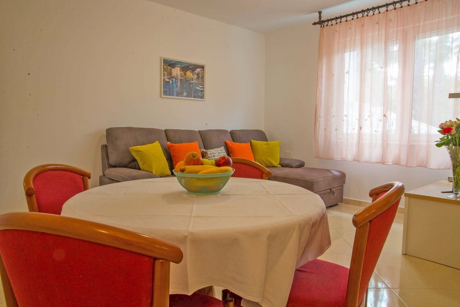 Ein Essbereich mit einem runden Tisch, Stühlen und einer Obstschale. Es gibt ein Sofa mit Kissen und ein Bild an der Wand.