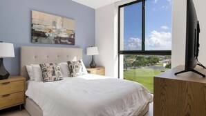 Ein Schlafzimmer mit einem großen Bett, zwei Nachttischen, einem Fernseher auf einem Schrank und einem Fenster mit Blick auf eine grüne Landschaft.