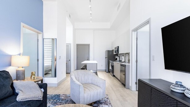 Ein modernes Wohnzimmer mit Sofa, Sessel, Couchtisch und Fernseher. Es gibt eine Küchenzeile mit Kühlschrank, Mikrowelle und Herd.
