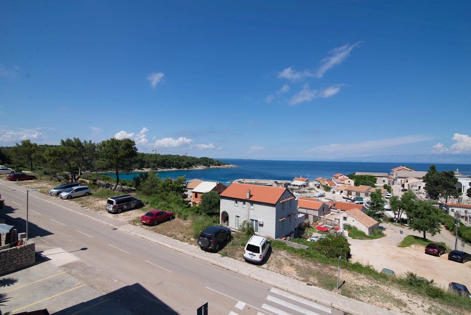 A coastal village with a road, parked cars, and buildings with red-tiled roofs.