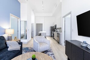 Un salon moderne comprenant un canapé, un fauteuil, une table basse et un téléviseur. Il y a également un coin cuisine équipé d’un réfrigérateur, d’un four à micro-ondes et d’une cuisinière.
