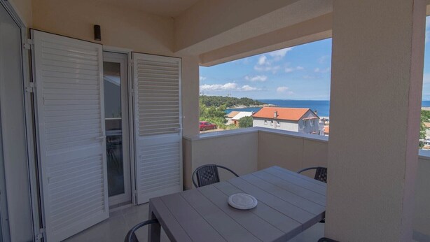 Un balcon avec une table et des chaises, une porte-fenêtre coulissante et une vue sur la mer et les bâtiments.