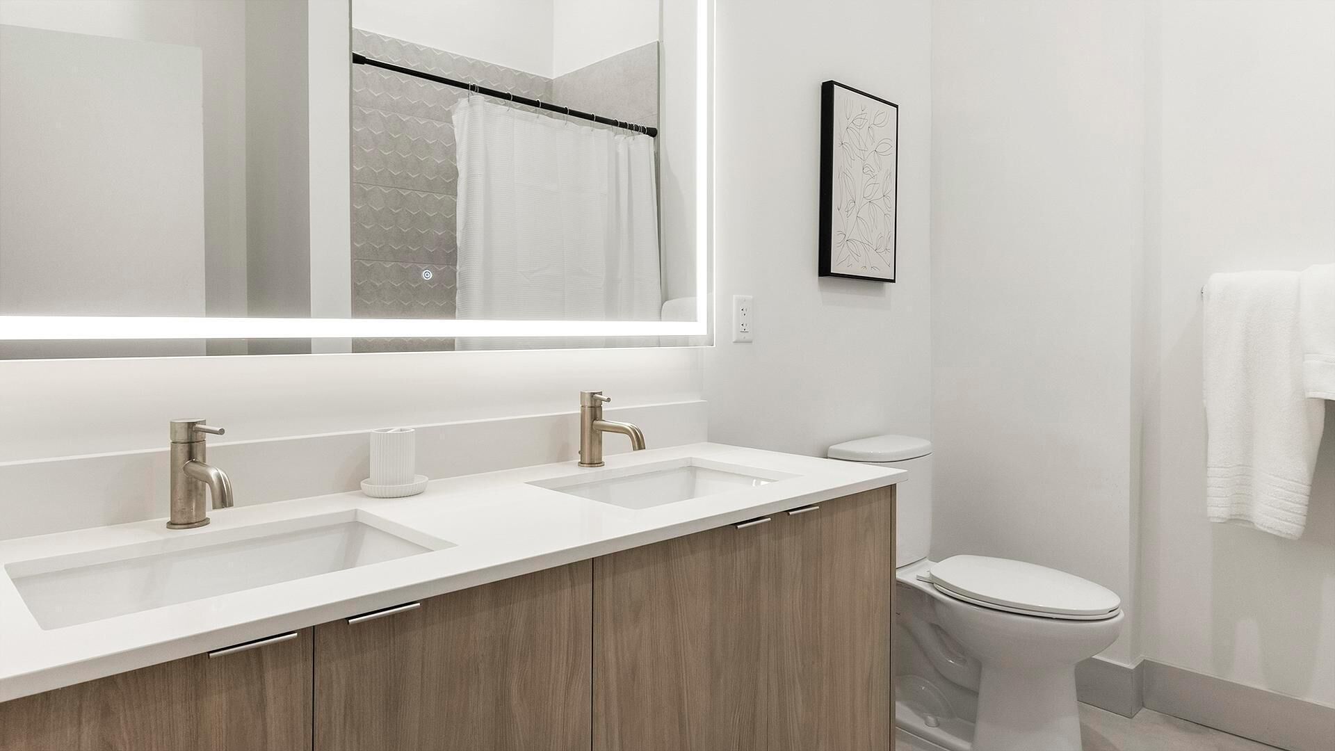A modern bathroom with a large mirror, two sinks, a toilet, and a shower curtain.