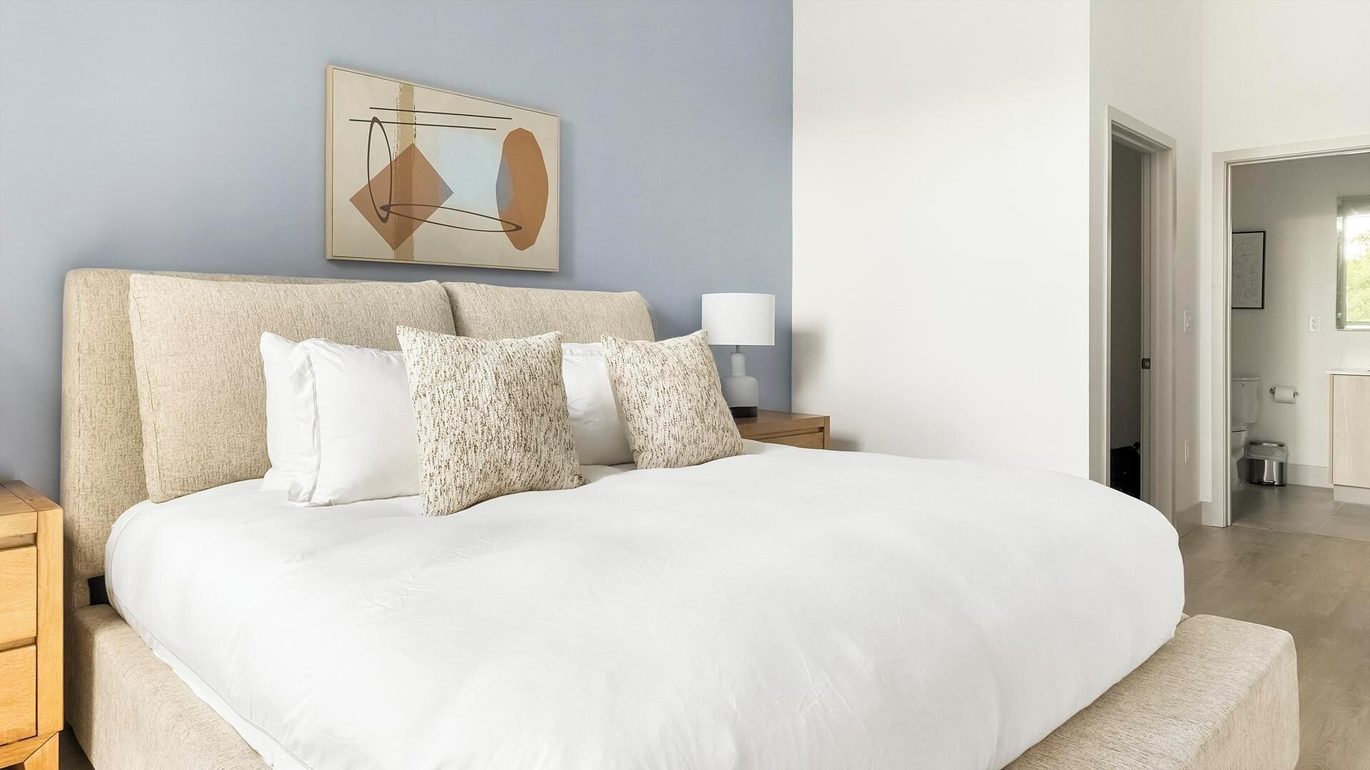 A neatly made bed with a white comforter and several pillows, a bedside table with a lamp, and a wall art piece above the bed.