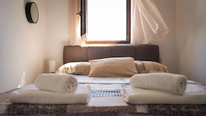 Una cama bien hecha con una colcha estampada, dos toallas enrolladas, una lámpara de noche y una ventana con cortinas.