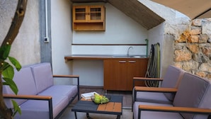 Una cocina exterior compacta con pileta y armarios de madera, rodeada por dos sofás morados y una pequeña mesa con fruta.