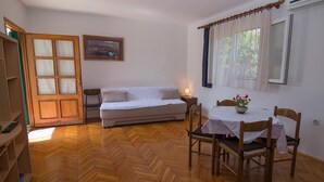 Een kamer met een houten visgraatvloer, een bank, een eettafel met stoelen, een raam met gordijnen en een deur.