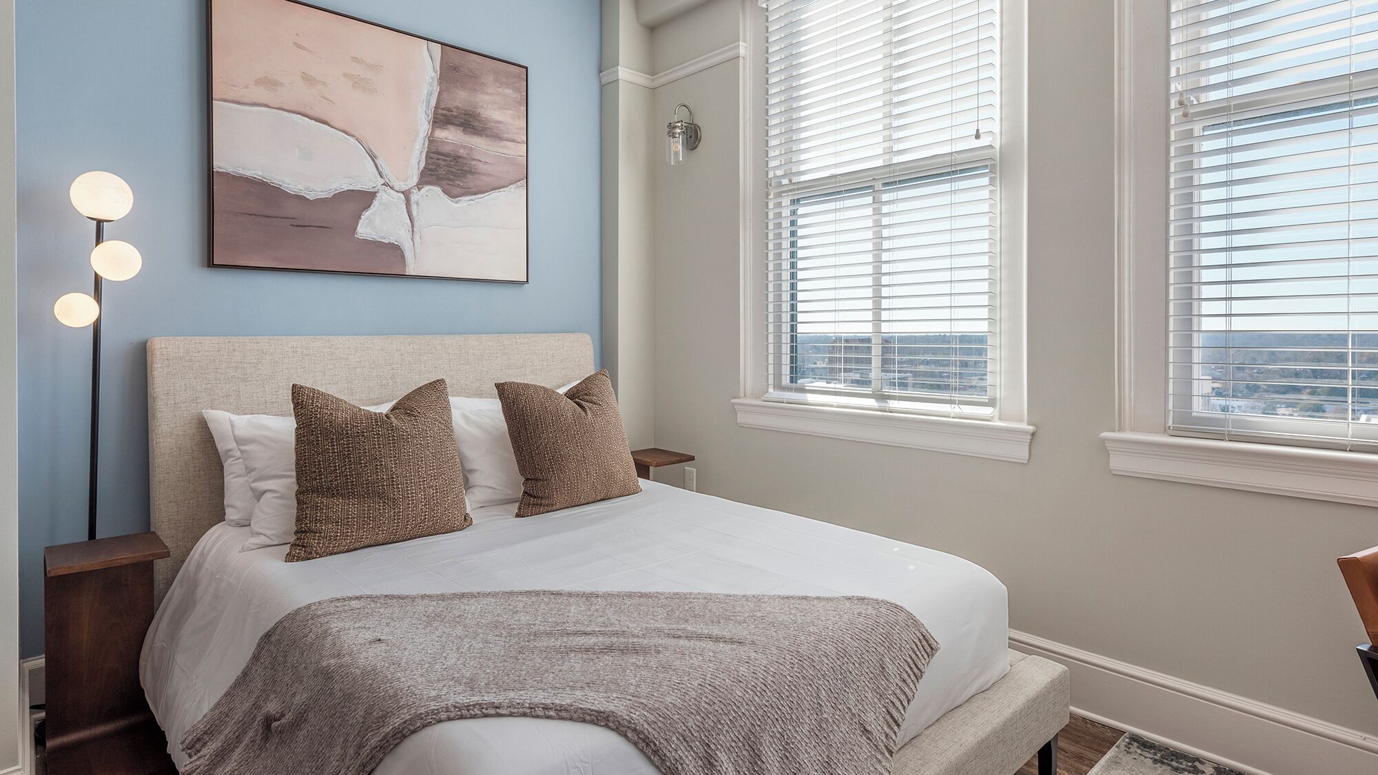 A bedroom with a bed, bedside lamps, a painting, and a window with blinds.