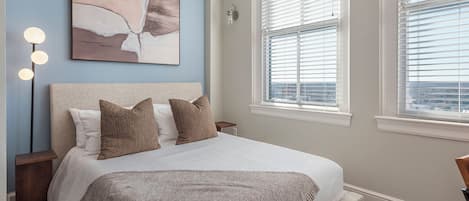 A bedroom with a bed, bedside lamps, a painting, and a window with blinds.