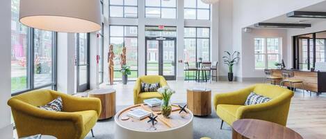 A spacious lobby with large windows, yellow armchairs, a central coffee table, and a reception desk.