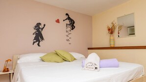 Ein ordentlich gemachtes Bett mit Kissen, einem zusammengerollten Handtuch und einem Paar Schuhe. Eine Wanddekoration zeigt eine Silhouette einer Person und ein Herz.