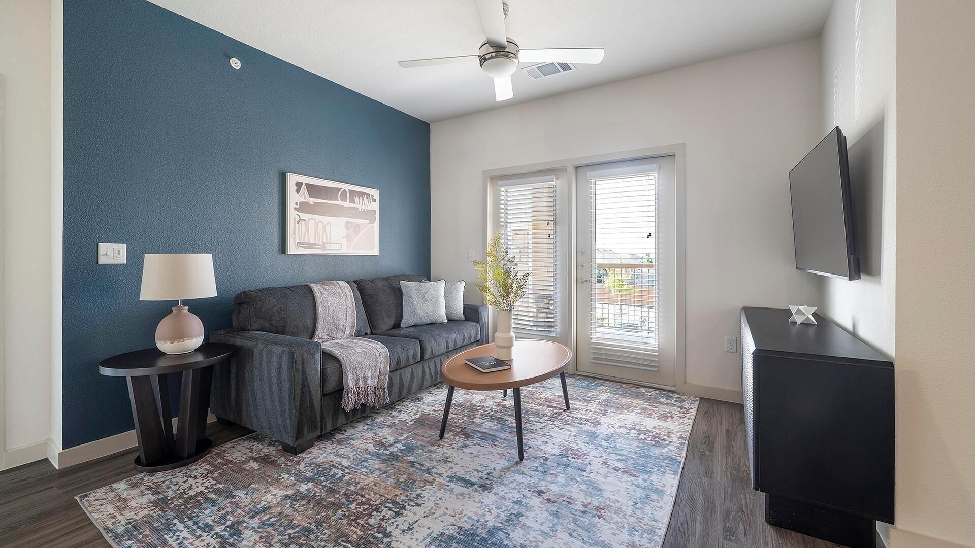 A modern living room with a blue accent wall, a grey sofa, a coffee table, a TV, and a ceiling fan.