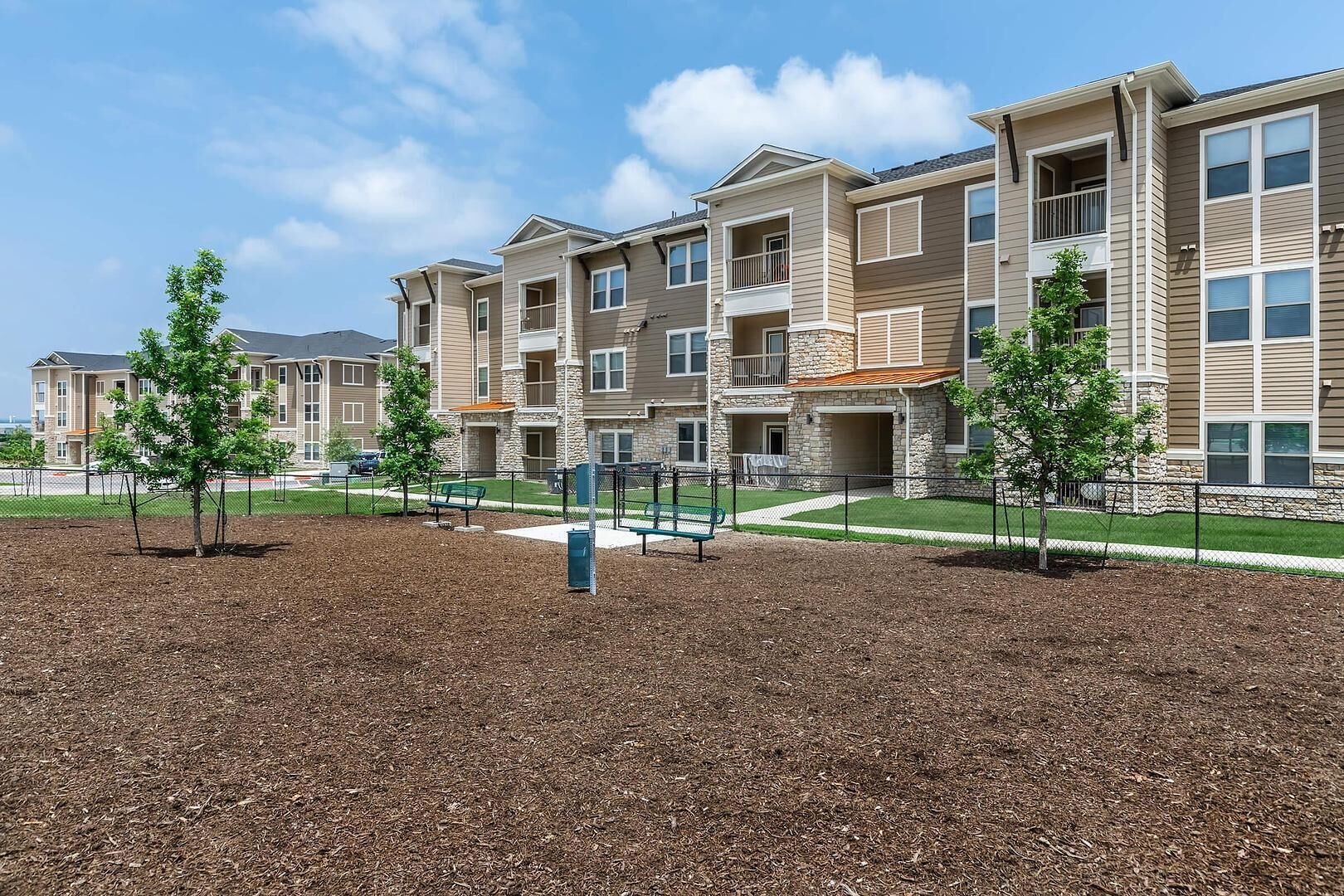 A multi-story residential building with balconies and a landscaped area with trees and a bench.