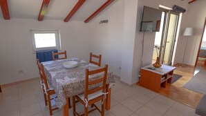 A dining area with a table set for four, wooden chairs, a TV mounted on the wall, and a window providing natural light. - Frantin / Frantin B4 (Mali Lošinj)