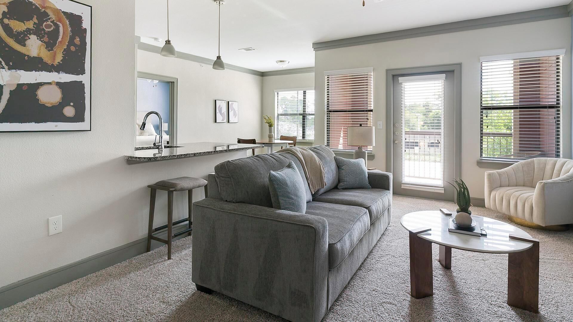 Una sala de estar moderna con un sofá gris, una mesa de centro redonda y un sillón blanco. Cuenta con una zona de cocina con un lavamanos y una ventana con persianas.