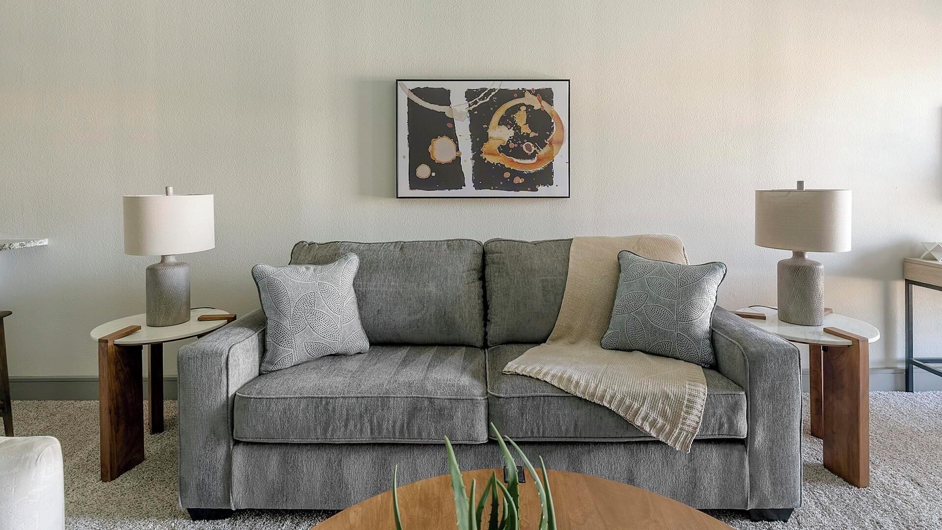 Una sala de estar con un sofá gris, dos mesitas de noche con lámparas, una obra de arte abstracta enmarcada y una planta pequeña sobre una mesa de centro de madera.
