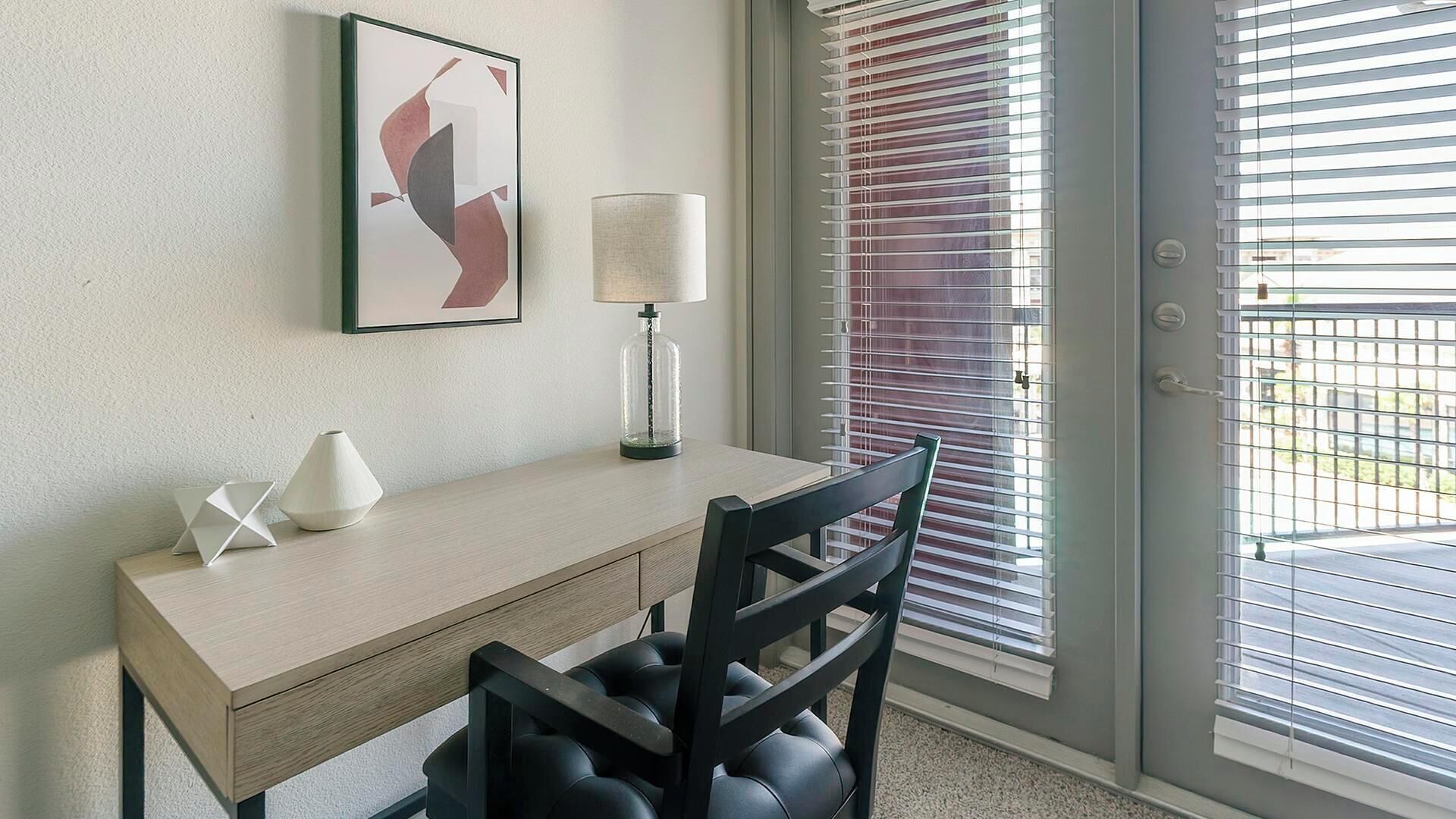 A hotel room with a wooden desk, a chair, a lamp, and a framed abstract artwork on the wall.
