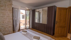 Un dormitorio con pared de piedra, cama con colcha estampada, armario de madera y puerta corredera de vidrio que da a un área exterior.