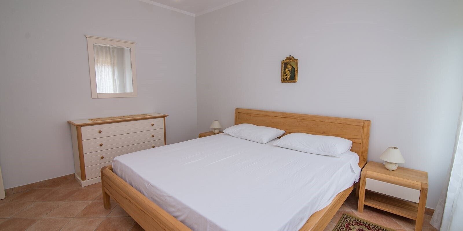 Een slaapkamer met een houten bed, twee nachtkastjes met lampen, een houten dressoir, een spiegel en een klein religieus beeld aan de muur.