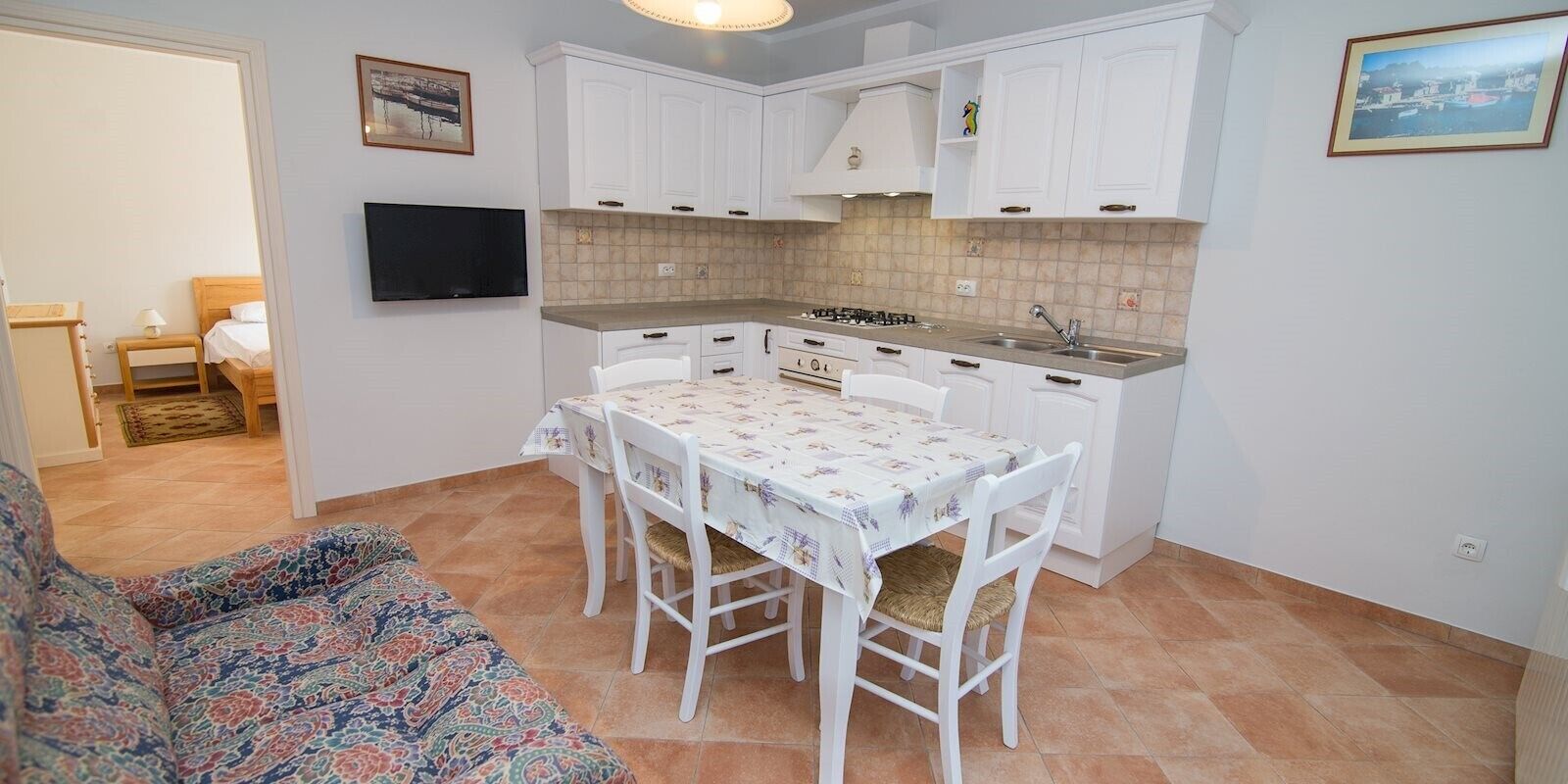 Una cocina compacta con armarios blancos, una mesa de comedor con sillas y un televisor montado en la pared.