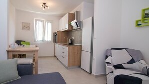 A compact kitchen with a wooden countertop, a built-in oven, and a white refrigerator. There is a window with curtains, a small table with a vase, and a sofa with a geometric pattern. - Ivana C6 in Veli Lošinj (Veli Lošinj)
