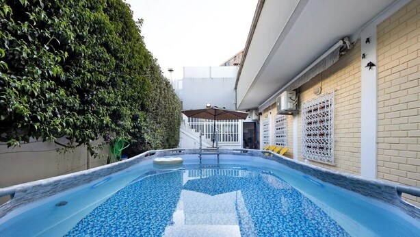A swimming pool with a blue mosaic floor, surrounded by a hedge and a building with a white exterior and a brick wall.