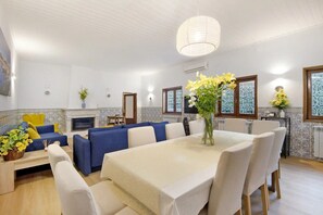 Zona pranzo con tavolo imbandito per sei persone, divani blu, camino e fiori decorativi.