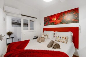 Una camera da letto con una testiera rossa, biancheria da letto bianca e un quadro con fiori rossi al muro.