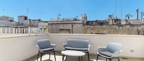 Une terrasse sur le toit, avec trois chaises grises et une petite table ronde, d’où l’on peut admirer un panorama urbain avec ses bâtiments et ses antennes.
