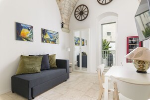 A living room with a dark sofa, a white table, and artwork on the walls. There is a door leading to another room and a balcony with a railing.