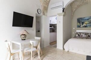 A bedroom with a bed, a TV, a desk with chairs, and a kitchen area in the background.