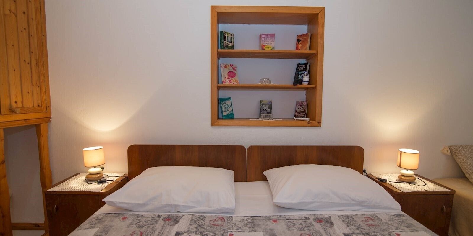 Un lit double avec deux oreillers, une tête de lit en bois et une table de chevet avec une lampe. Au-dessus du lit, on trouve une étagère avec des livres et un petit objet décoratif.