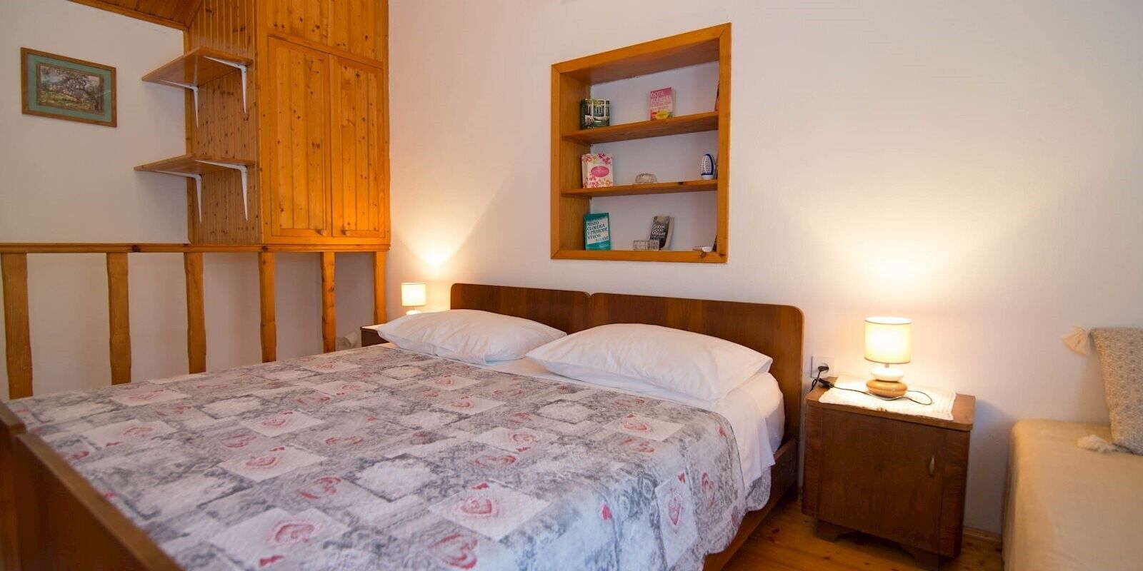 A bedroom with a wooden bed, a nightstand with a lamp, a shelf with toiletries, and a wooden wardrobe.