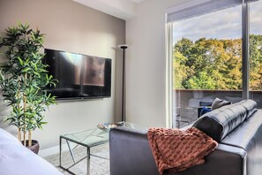 A modern living room with a flat-screen TV, a glass coffee table, a sofa with a throw blanket, and a large window offering a view of trees. - Landing | Incredible Studio, Gym (Ithaca)