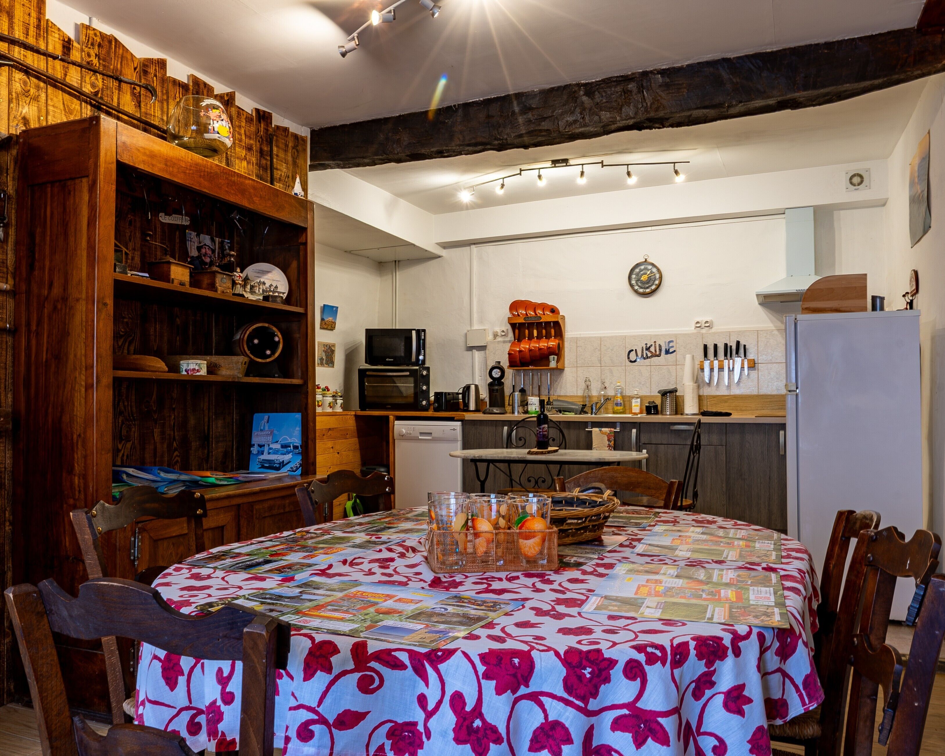 Een keuken met houten planken, een eettafel met een tafelkleed met bloemenmotieven en een klok aan de muur.