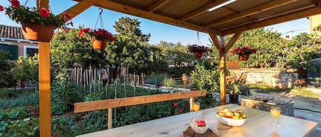 An outdoor patio with a wooden table, hanging flower pots, and a stone wall in the background.