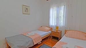 Une petite chambre propre, avec deux lits simples, une table de chevet avec une lampe et une fenêtre ornée de rideaux légers.