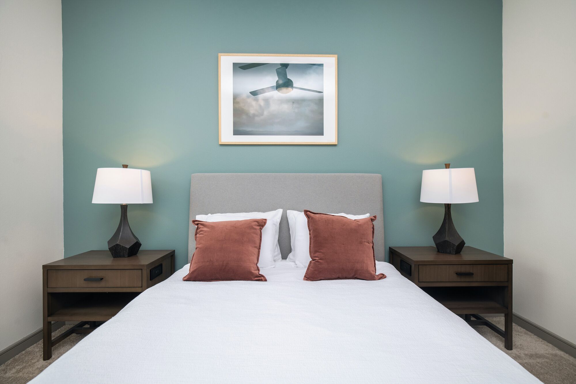 A neatly made bed with two pillows, a framed picture above, and two bedside tables with lamps.