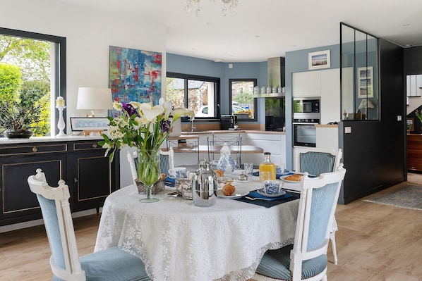 A dining area with a table set for breakfast, a kitchen in the background, and a view of the outdoors.