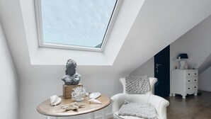 A room with a skylight, a white chair, a small wooden table with decorative items, and a white dresser.