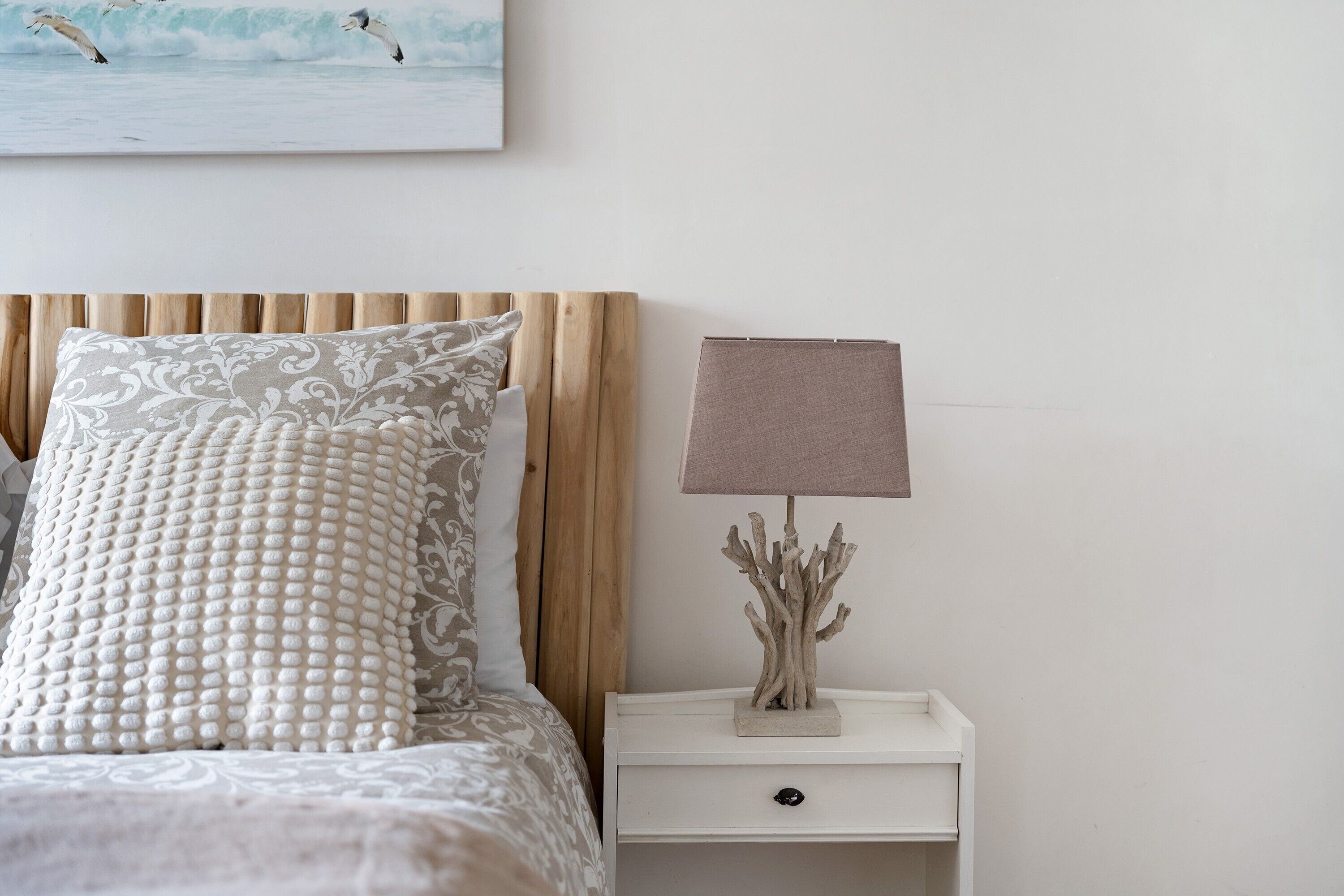 Un letto rifatto con cuscini decorati, una testiera in legno, un comodino bianco con una lampada e un oggetto decorativo, e un quadro appeso al muro.