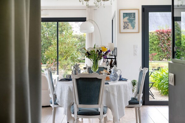 Ein Essbereich mit einem Tisch für vier Personen, Stühlen mit blauen Kissen, einer Vase mit Blumen und Blick ins Grüne.