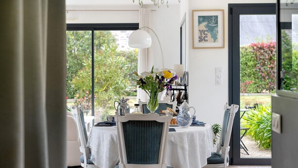 Ein Essbereich mit einem Tisch für vier Personen, Stühlen mit blauen Kissen, einer Vase mit Blumen und Blick ins Grüne.