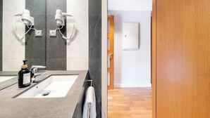 Een moderne badkamer met een grote spiegel, twee haardrogen, een wastafel met een zeepdispenser en een handdoekhouder.