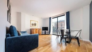 A modern living room with a blue sofa, a dining table with chairs, a TV, and a large window with curtains. - Spacious 1 bedroom apartment with parking in Eixample (Barcelona)