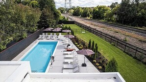 En pool på taket med solstolar och parasoller, omgiven av en välskött gräsmatta och med ett järnvägsspår synligt i bakgrunden.