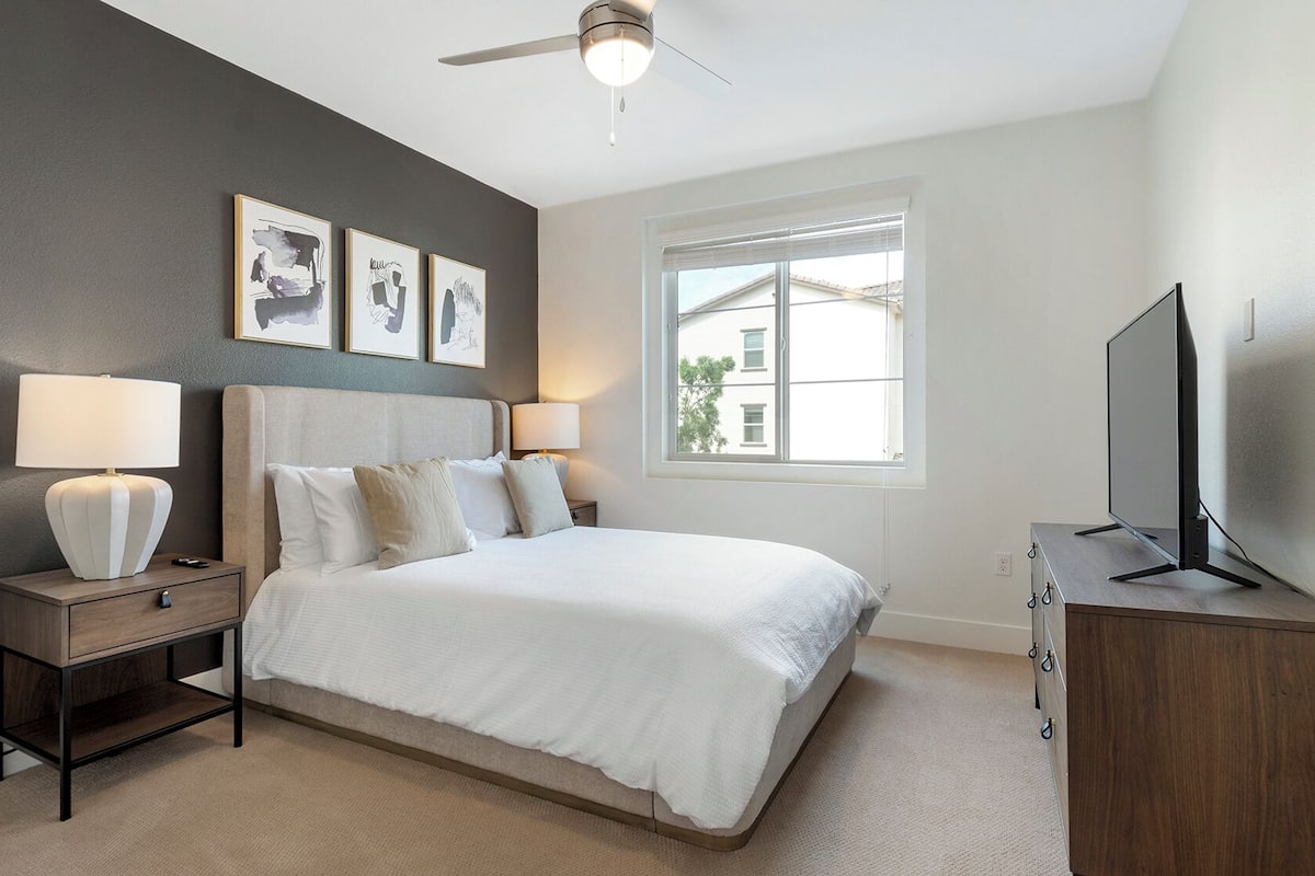 A bedroom with a bed, two bedside tables with lamps, a ceiling fan, a window with blinds, and a TV on a wooden cabinet.
