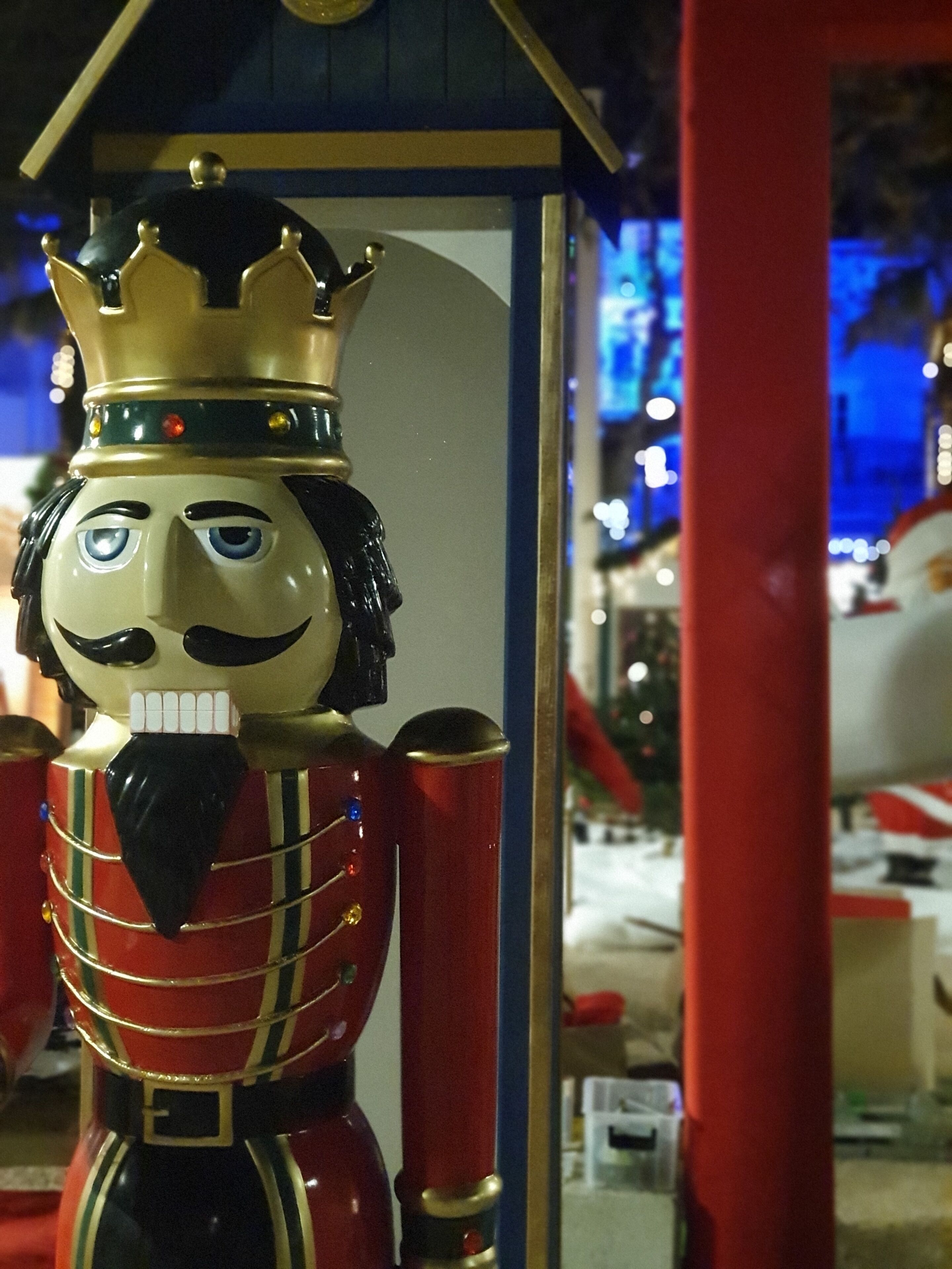 A nutcracker figure with a crown and a mustache, standing in front of a building with a blue roof.