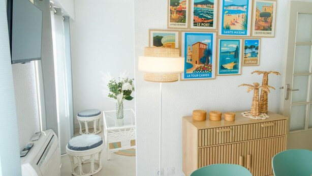 Ein Zimmer mit weißer Wand, einem Holzkabinett mit Dekorationsgegenständen, einer Lampe und einer Reihe gerahmter Poster mit verschiedenen französischen Orten.