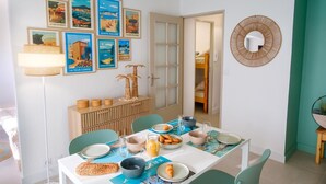Ein Essbereich mit einem Tisch, gedeckt für vier Personen, mit verschiedenen Speisen und Getränken. An der Wand über dem Tisch hängen gerahmte Bilder, und auf der Anrichte steht ein Brotkorb.